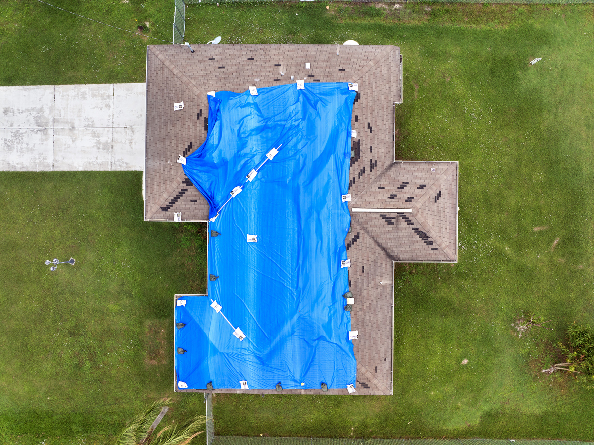 Hurricane affected house roof with blue tarp stretched over broken area to guard against rain until full asphalt shingle restoration can be carried out.