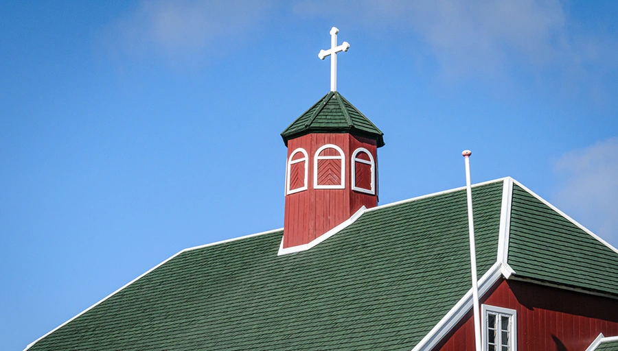 Green shingle roof on a red church building with a steeple and cross against a blue sky