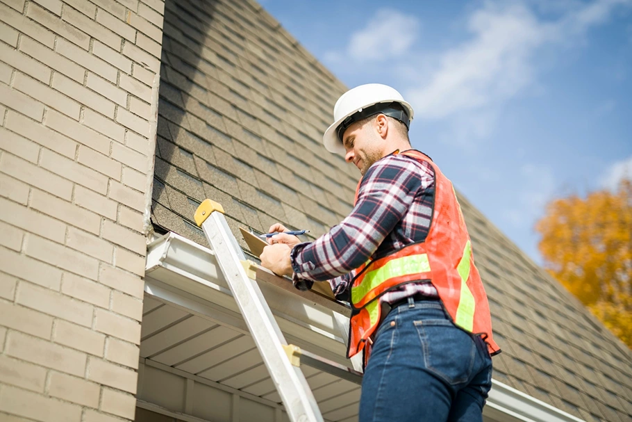 Roofing inspector on a ladder documenting shingle roof and gutter damage on a clipboard
